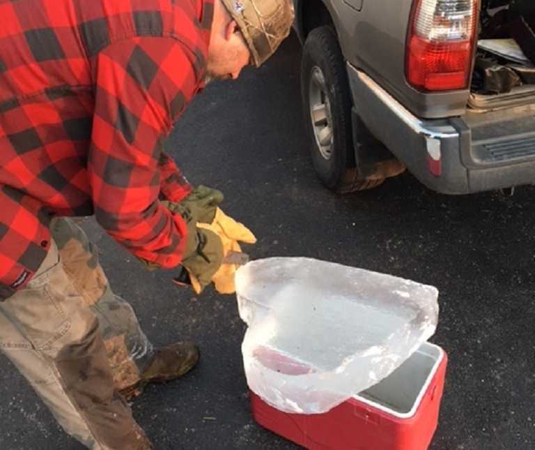man with knife carves piece of ice resting on cooler into a heart next to his car with trunk open 