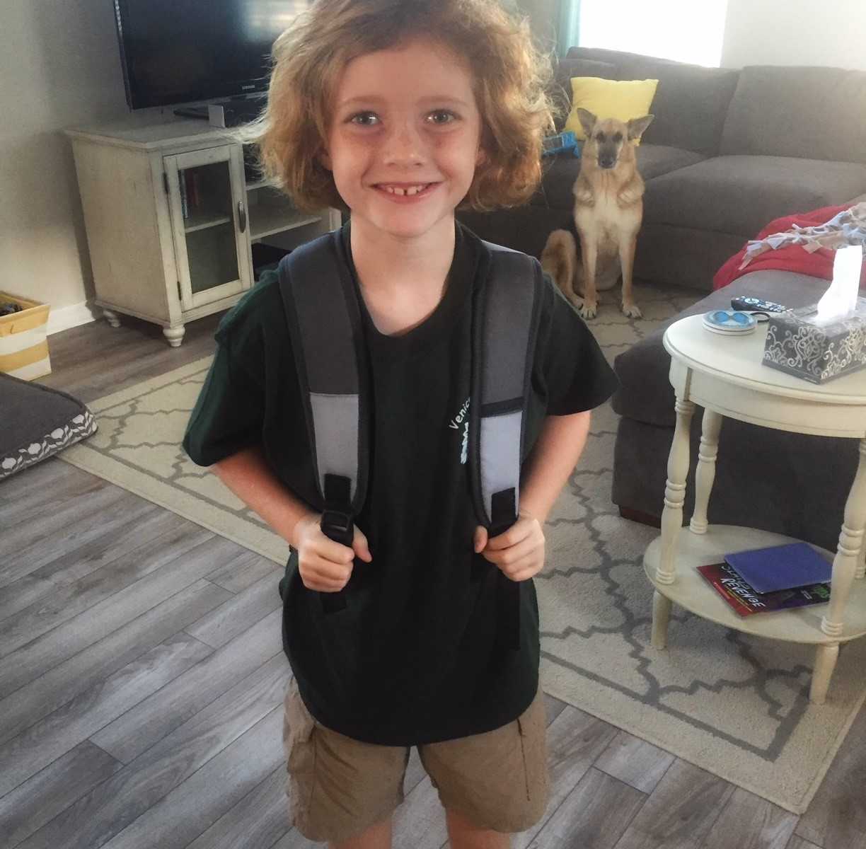 young boy standing in living room smiling with back pack on and german shepherd in background