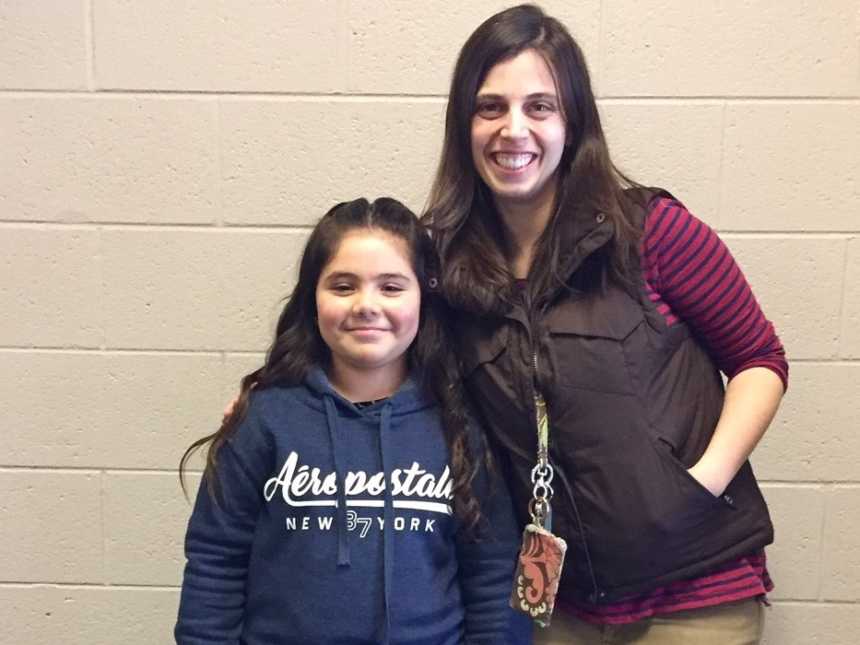 young girl in navy sweatshirt stands next to woman with hand in pocket of vest smiling in front of cinderblock wall