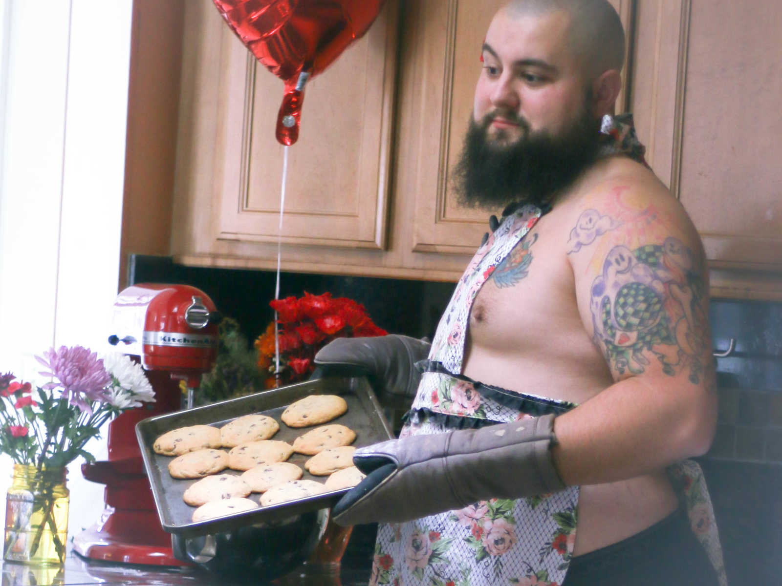 shirtless man wearing apron holds baking sheet of chocolate chip cookies in kitchen with flowers and heart balloon