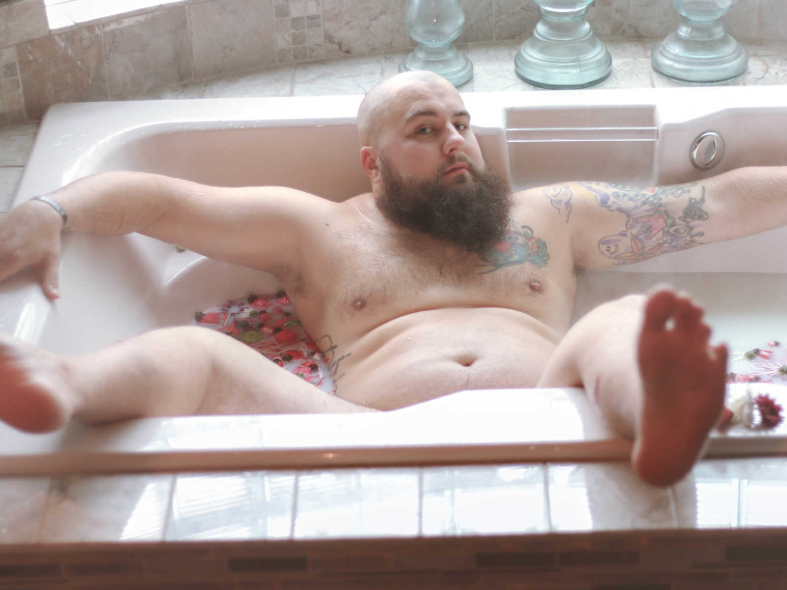 man poses in bathtub with flowers in the water with him