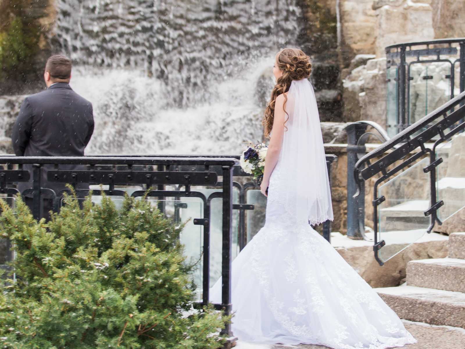 bride in wedding dress staring at back of the groom who is standing on a bridge next to a waterfall