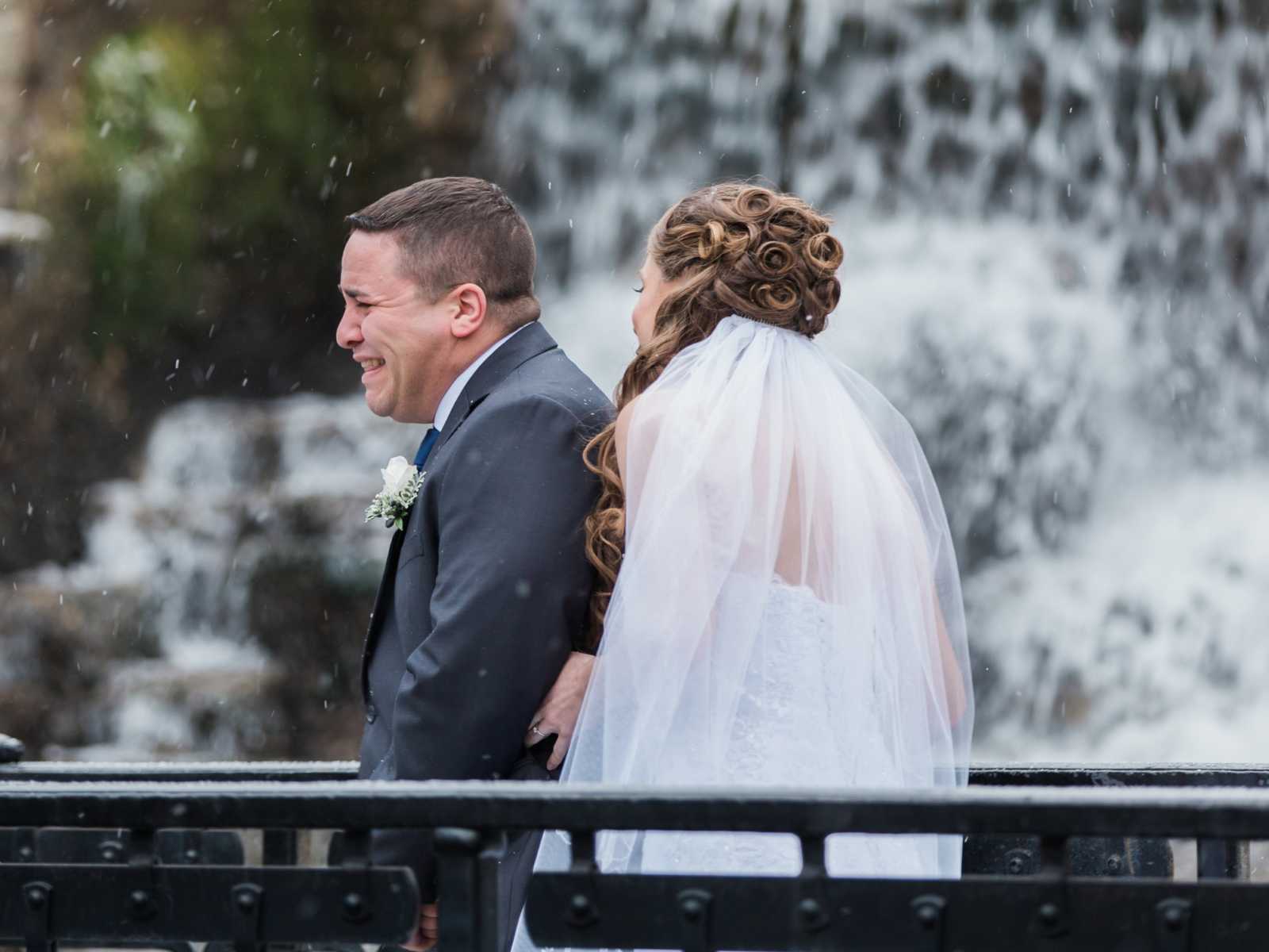 bride holds groom from behind as groom is in tears