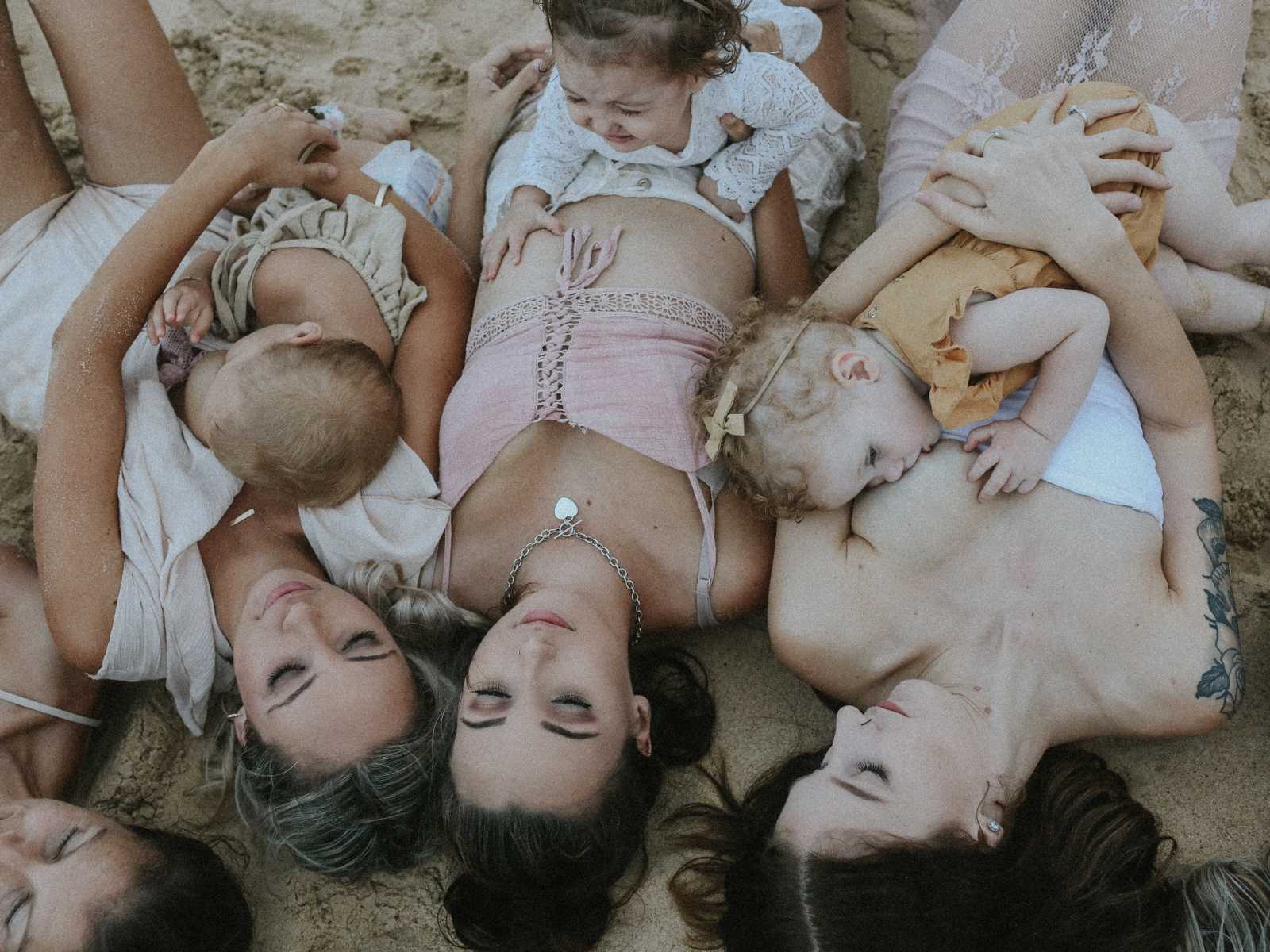 Woman lie in a circle with their heads together while breastfeeding on the beach