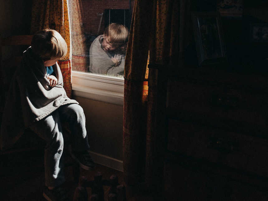 Little boy sits in dark room with blanker wrapped around him looking at his reflection in the window