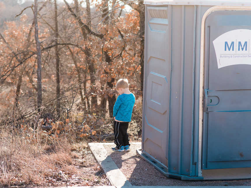Little boy going to the bathroom in the woods beside a port-a-potty
