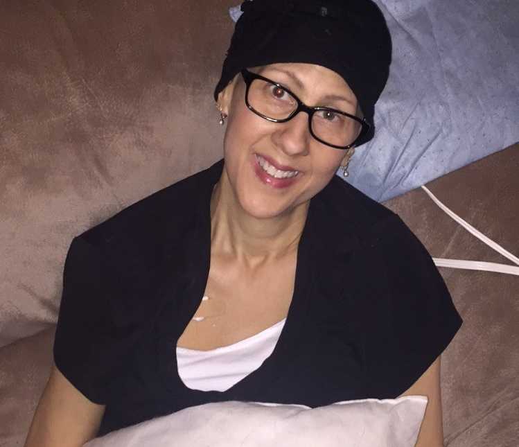 Mother with cancer who has since passed sits in chair looks up smiling