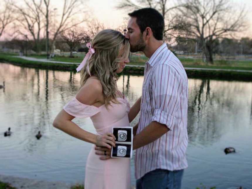 Husband kisses forehead of pregnant wife as she holds ultrasound picture in front of pond