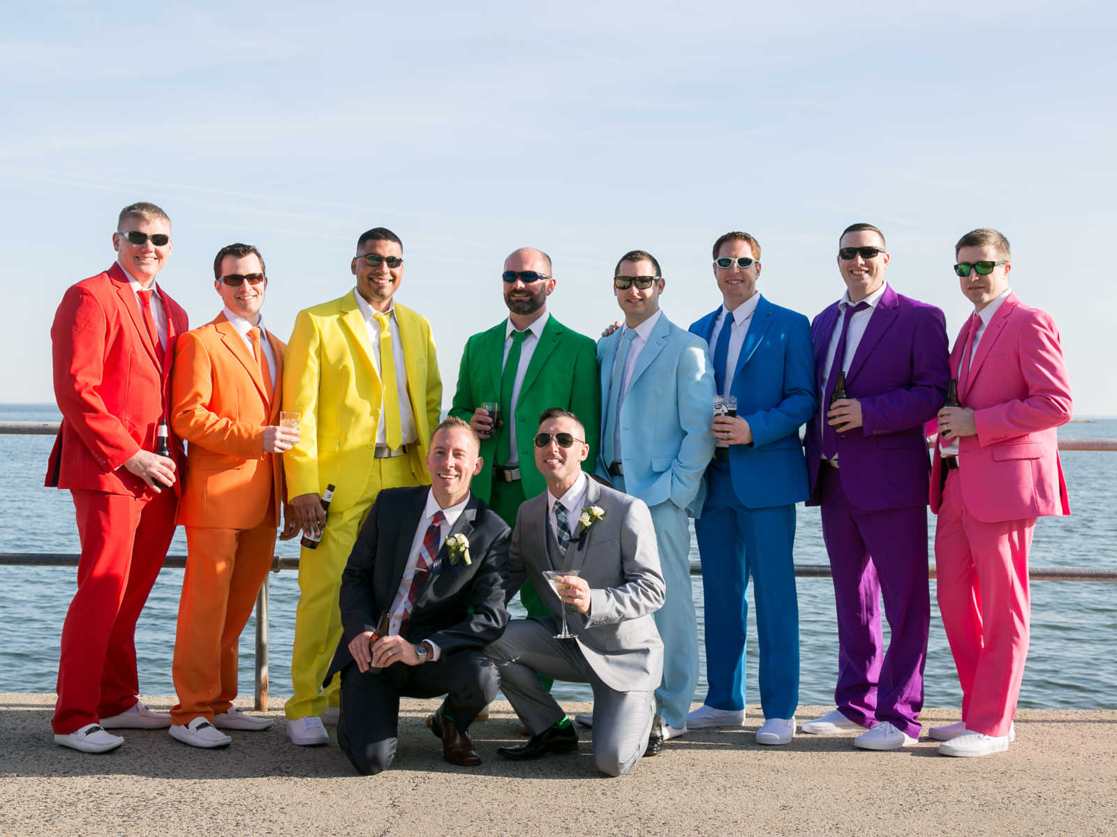 Grooms kneel on pier smiling in front of friends who are lined up in rainbow colored suits
