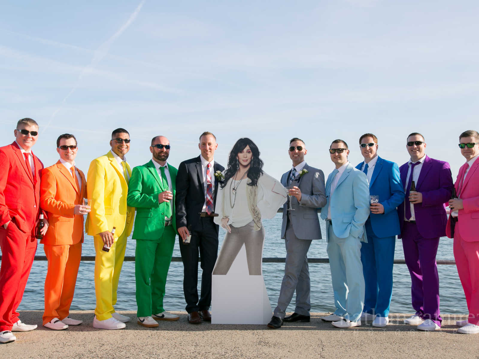 Grooms standing on pier with cutout of Celine Dion in between them and men in colorful suits on either side of them