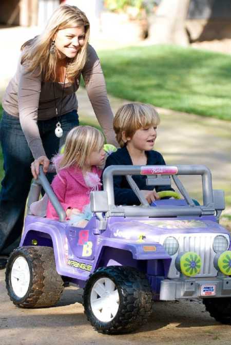 Mom smliles while pushing son and daughter with special needs in electric toy jeep 