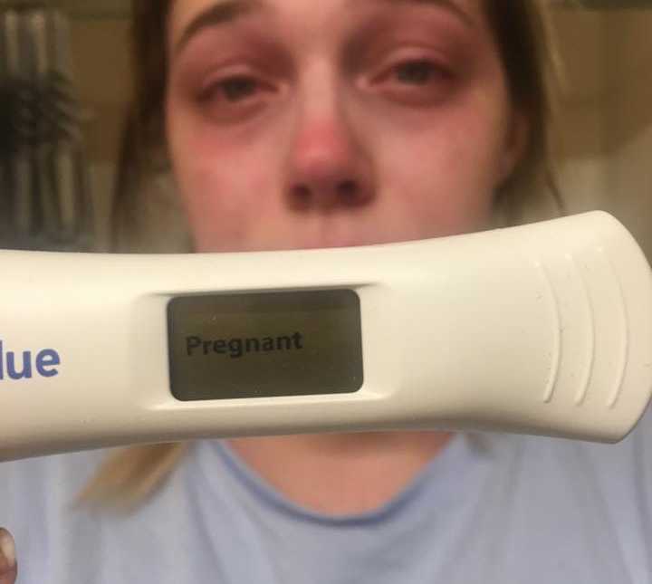 Selfie of woman holding up pregnancy test close to the camera that says, "pregnant" while she cries in background