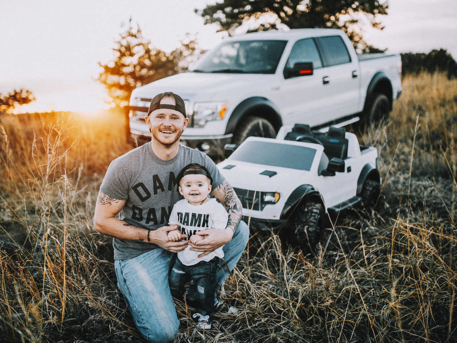 Father neals in grass with toddler in matching outfits in front of toy truck and real truck