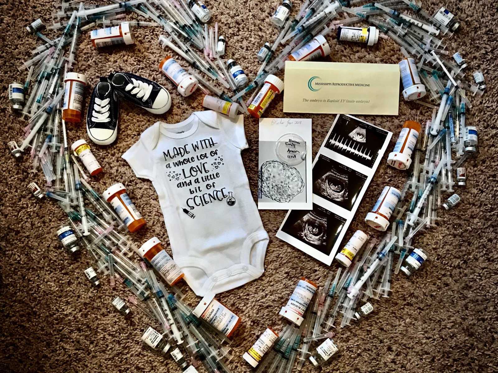 IVF needles and medication in shape of heart enclosing baby onesie, baby shoes, and ultrasound