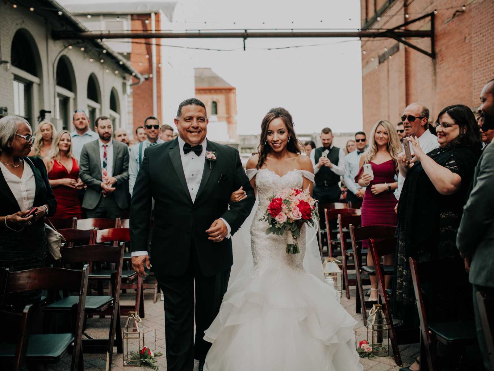 Father and daughter smile as father walks daughter down the aisle while crowd watches