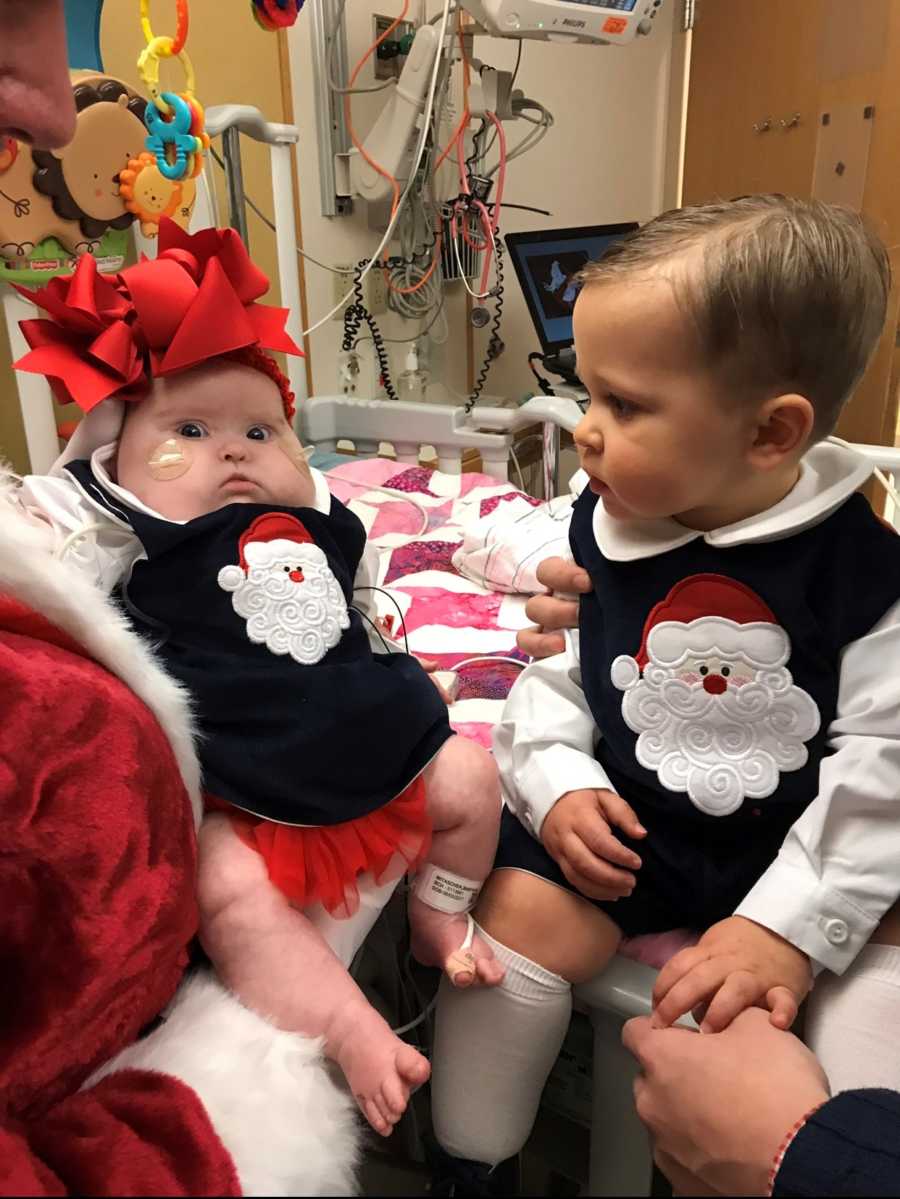 Infant brother in Santa onesie sits in hospital bed looking at baby sister with heart defect in same onesie
