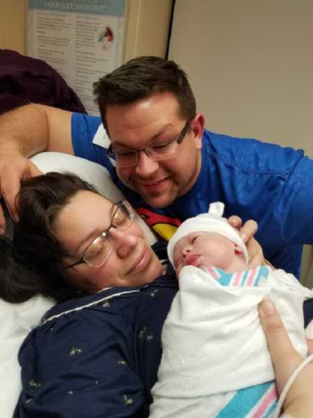 Mother lays in hospital bed with newborn who has down syndrome with husband over her shoulder