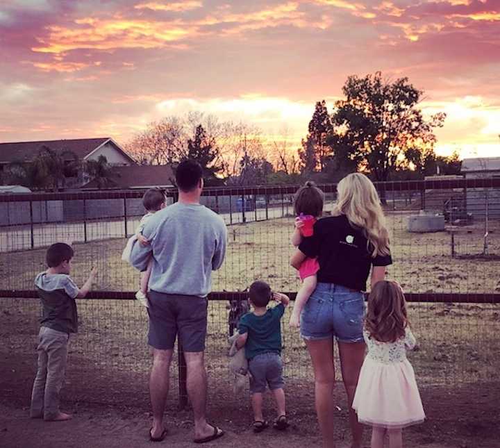 Mother and father holding two little girls with two young boys and girl standing at their side looking at farm animals