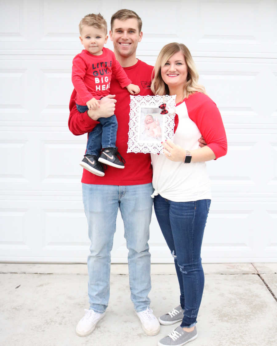 Father holds first born while wife stands next to him holding frame of their baby daughter who passed away