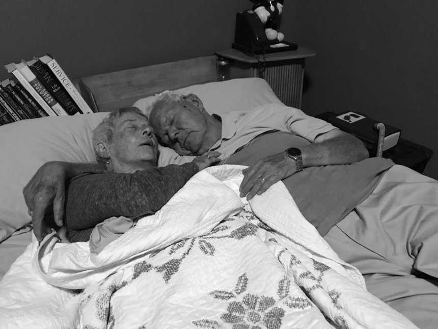 Husband has arm around wife with dementia at they lie asleep in bed