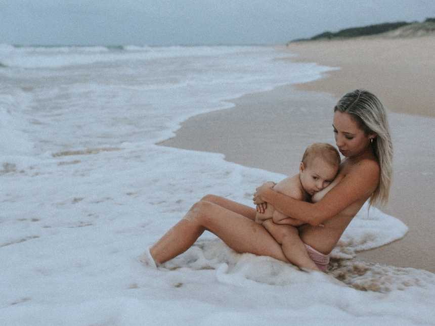 Naked woman breastfeeding on the beach as the tide comes in 