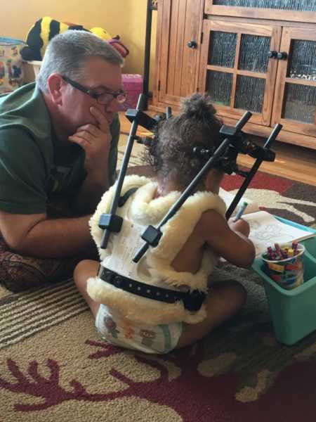 Toddler who was internally decapitated sits on floor at home coloring wearing a halo while dad watches