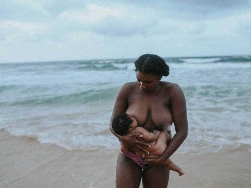 Woman stands with her back to the ocean as she breastfeeds