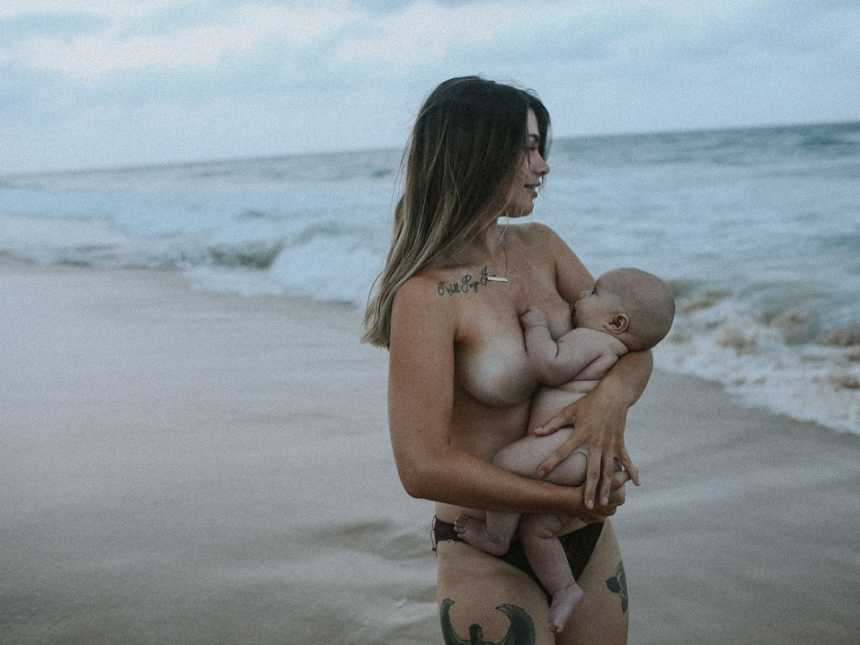 Topless woman stares out at the ocean while breastfeeding on the beach