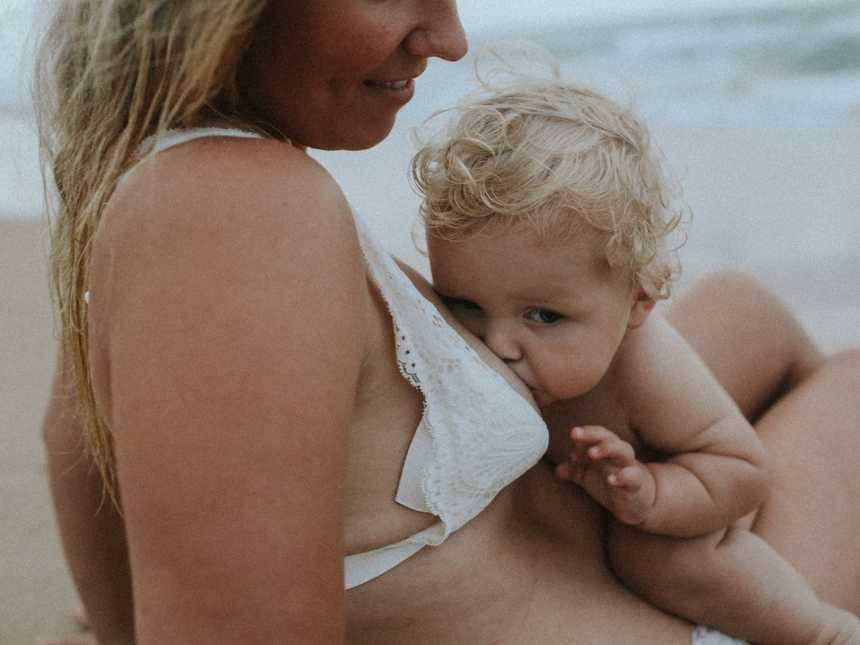 Woman looks down smiling at her baby who is breastfeeding on the beach