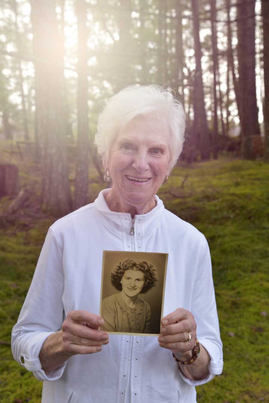 90 year old woman stands in the woods holding picture of younger self