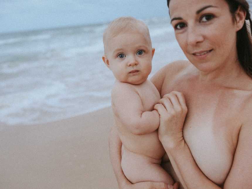 Topless woman holds her baby by the shore of the ocean