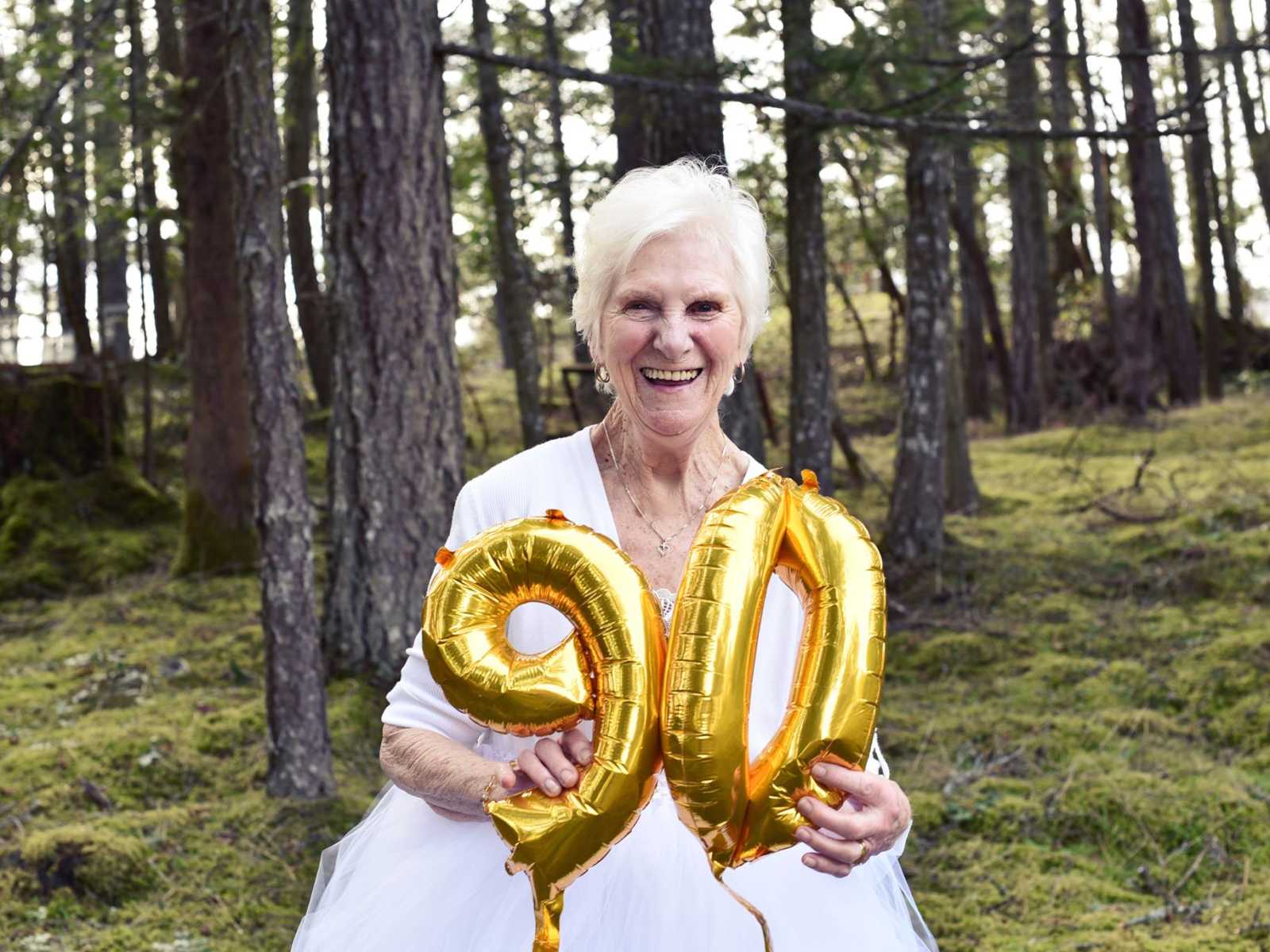 90 year old woman smiles in the woods while holding balloons that are number 9 and 0
