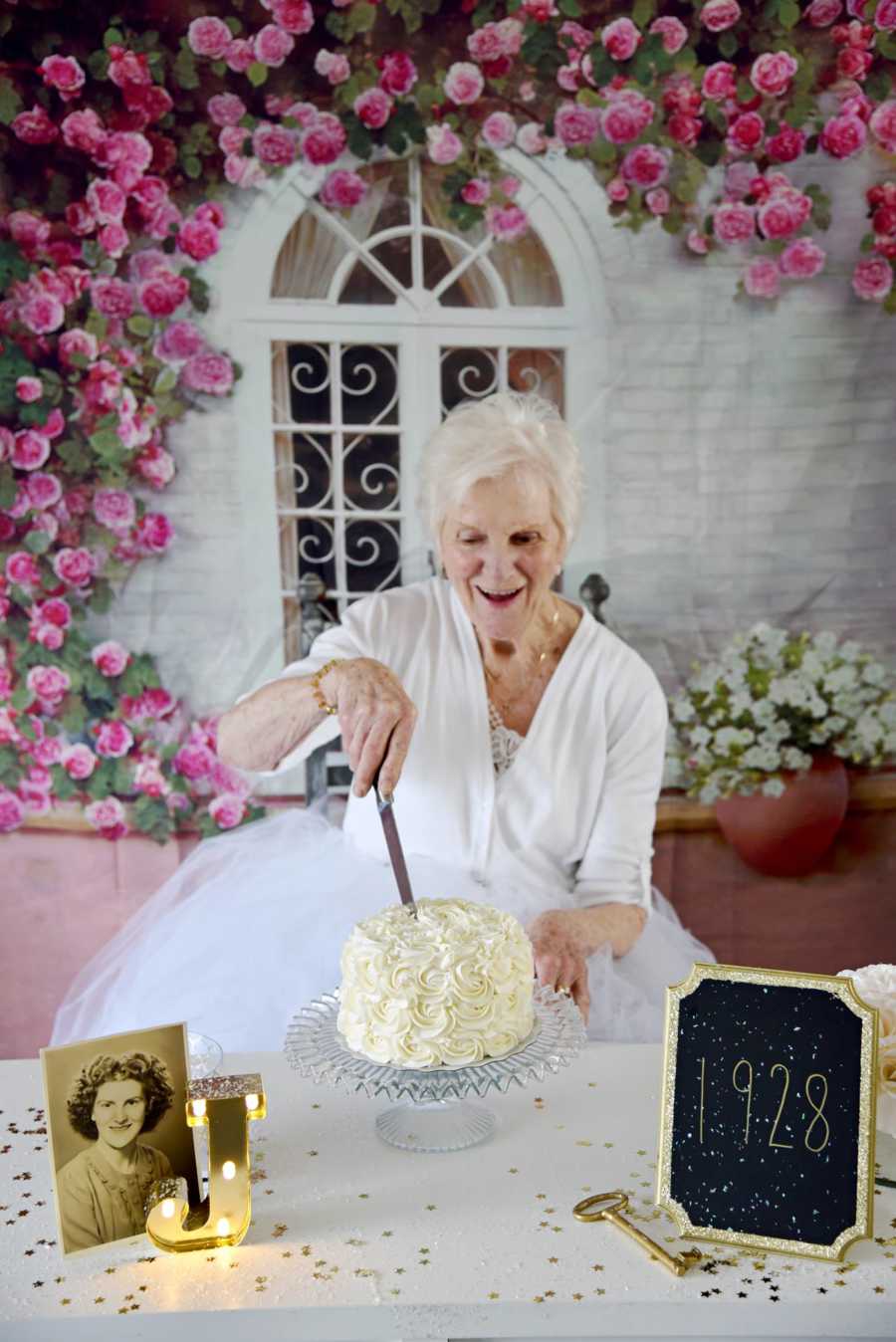 90 year old woman cuts in to birthday cake next to light up letter J, picture of her younger self and chalk board