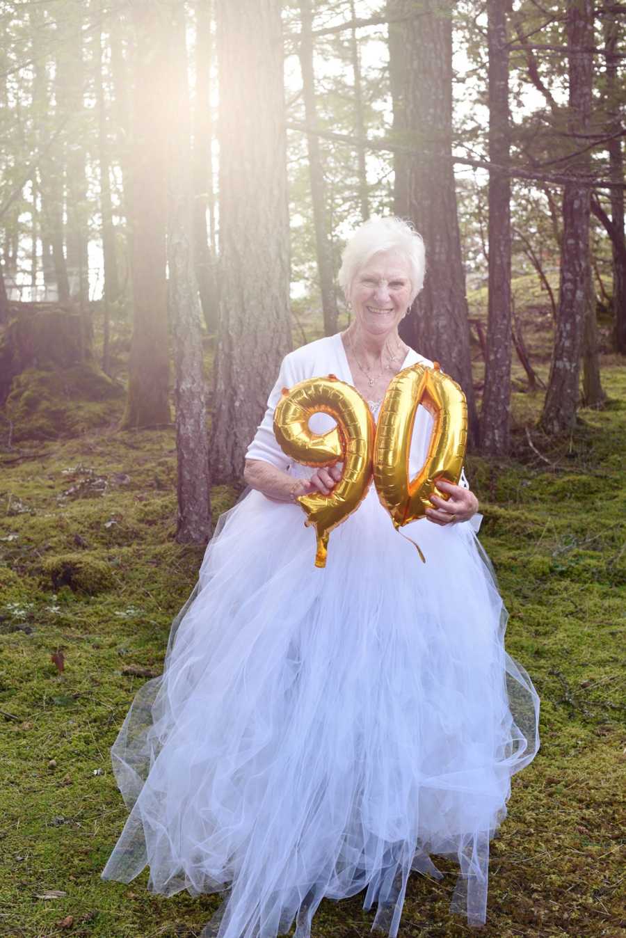 90 year old woman standing in woods in white dress holding up gold balloons that say 90