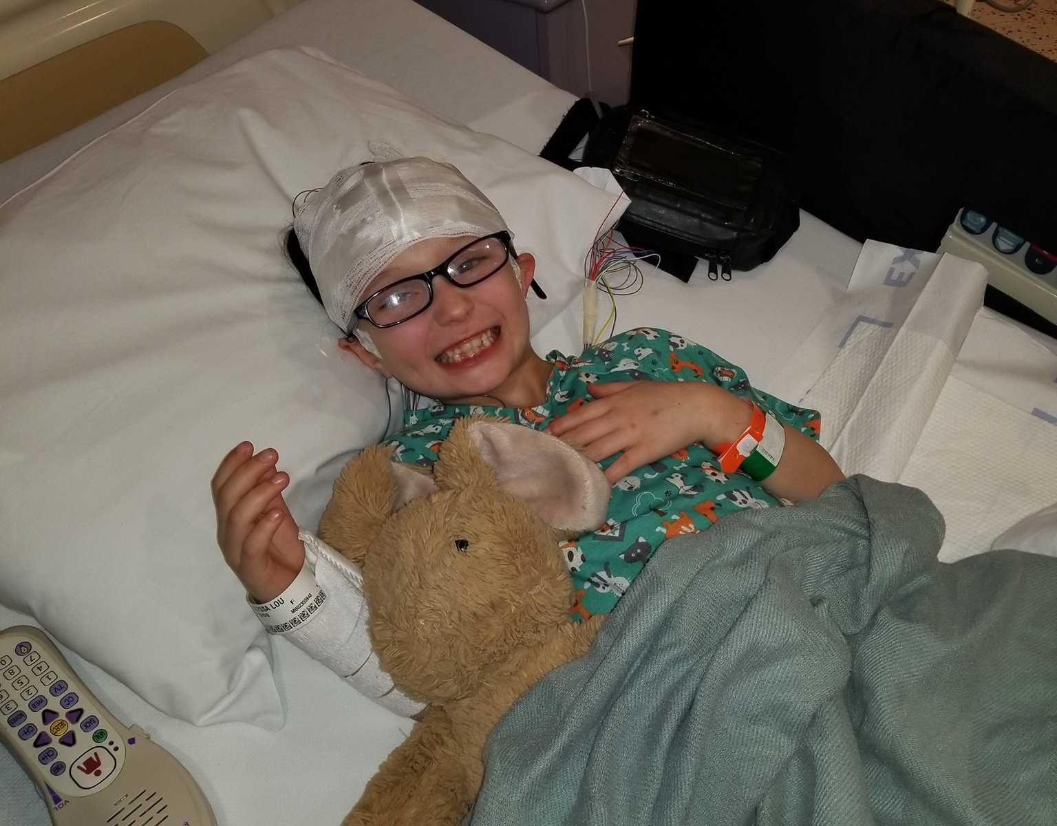 Nine year old who suffers from seizures smiling in hospital bed with stuffed animal and bandage on head