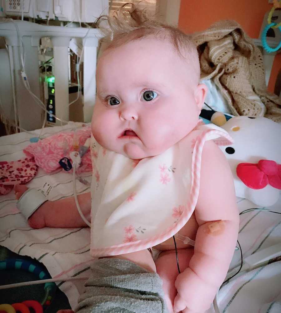 Baby girl with heart defect sits up in hospital bed with bib with pink flowers on it