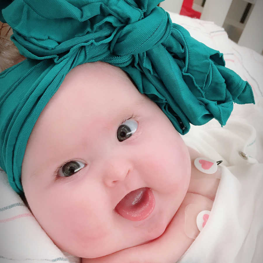 Baby with heart defect and low glucose levels smiles in hospital bed with large green bow on her head