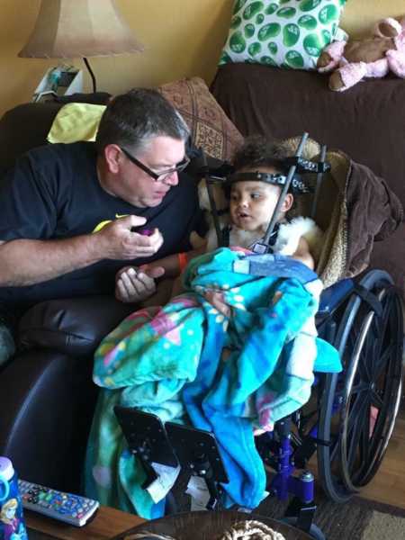 Toddler who was internally decapitated sits in wheel chair wearing a halo next to dad sitting in recliner