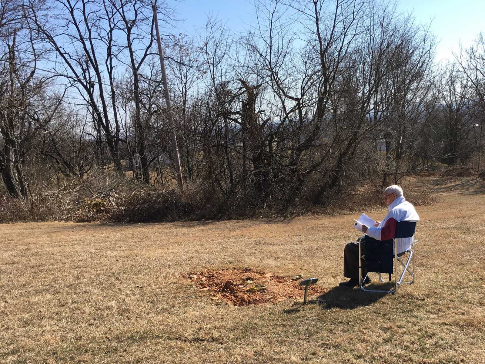 Husband of wife with dementia sits near grave of late son reading in a chair