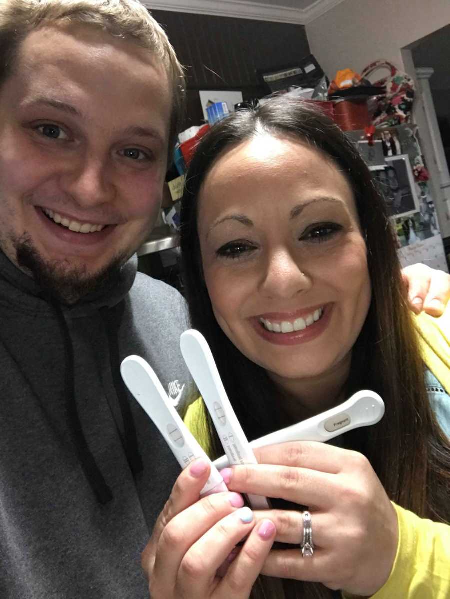 Husband and wife smile in selfie with three pregnancy tests that all say, "pregnant"