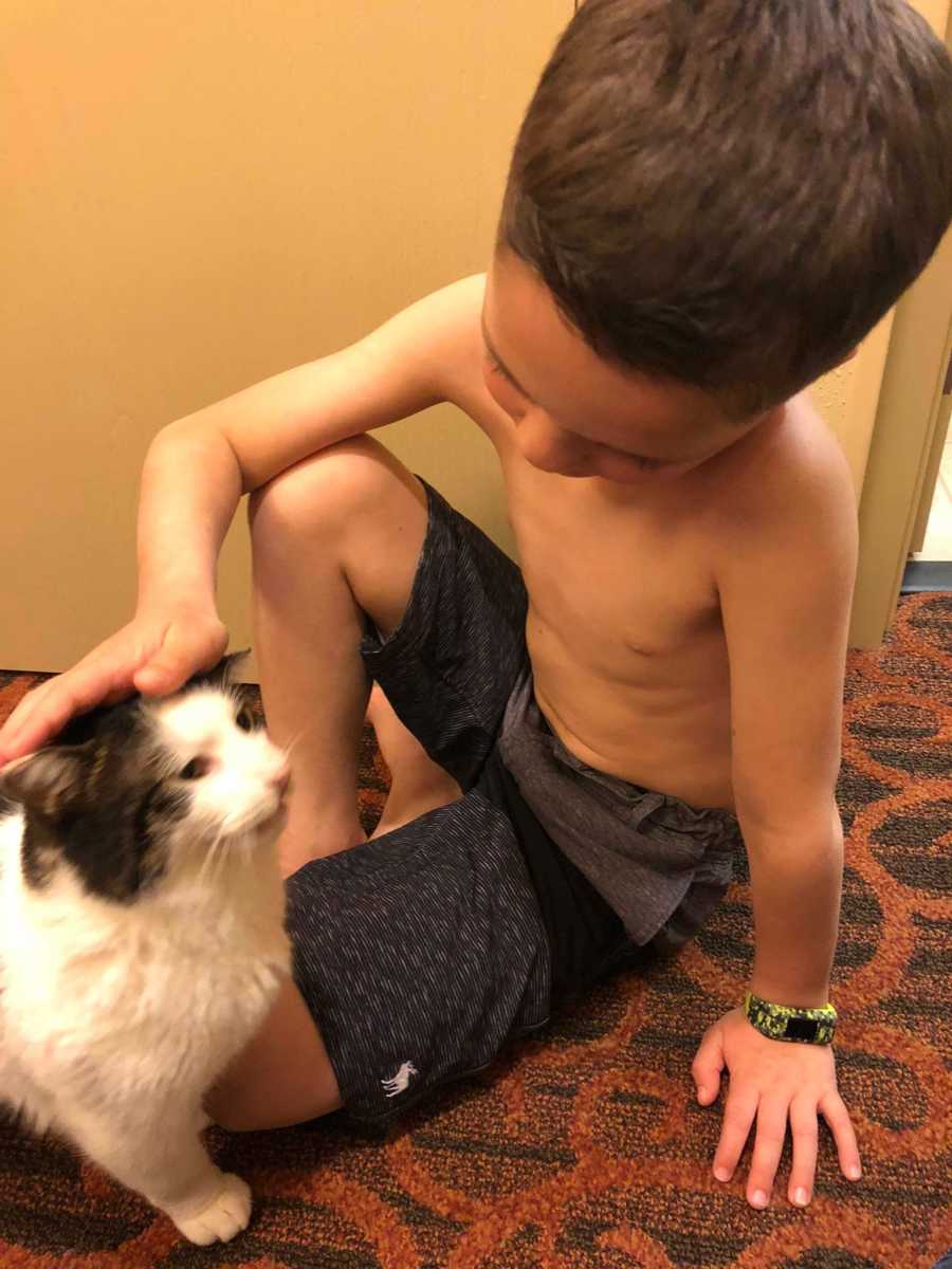 Boy with two different colored eyes sits shirtless on ground petting his rescue cat