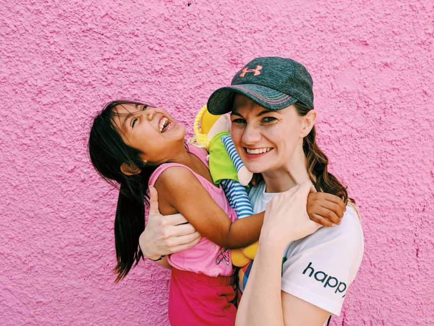 Woman smiling holding adopted daughter from Mexico in front of pink wall