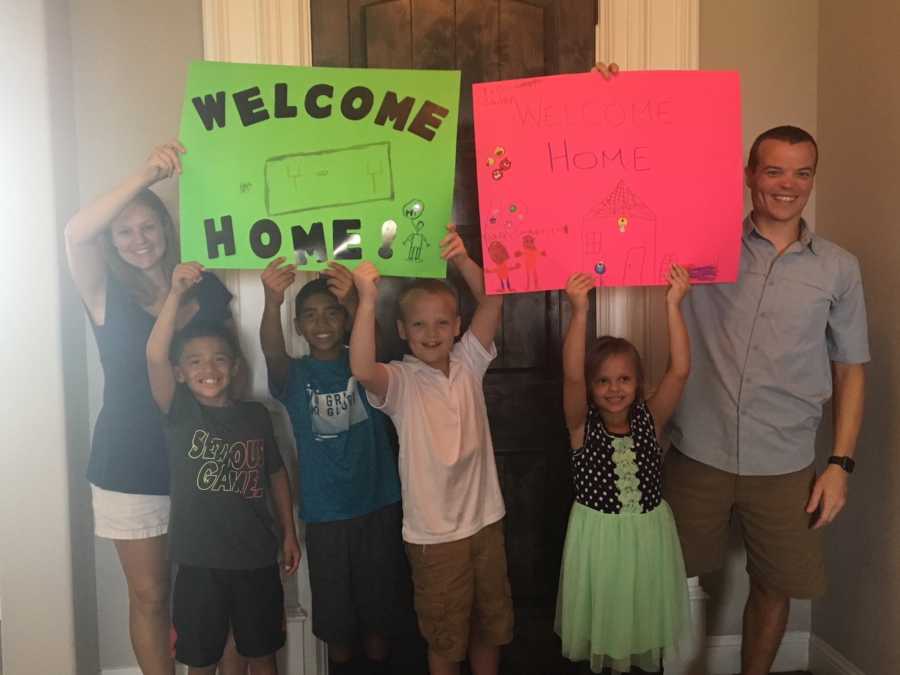 Mother and father along with their two birth children and two adopted children holding up posters saying, "welcome home"