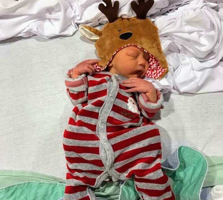 Miracle baby sleeping on bed wearing grey and red onesie with reindeer hood over his head
