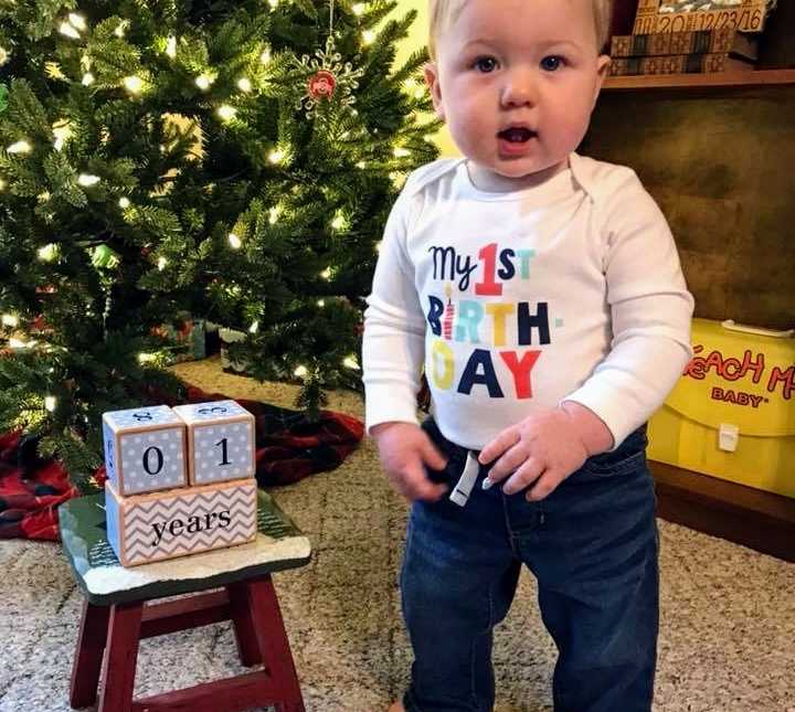 Miracle baby in onesie saying, "my 1st birthday" stands next to Christmas tree and stool with wooden blocks saying, “1 years”