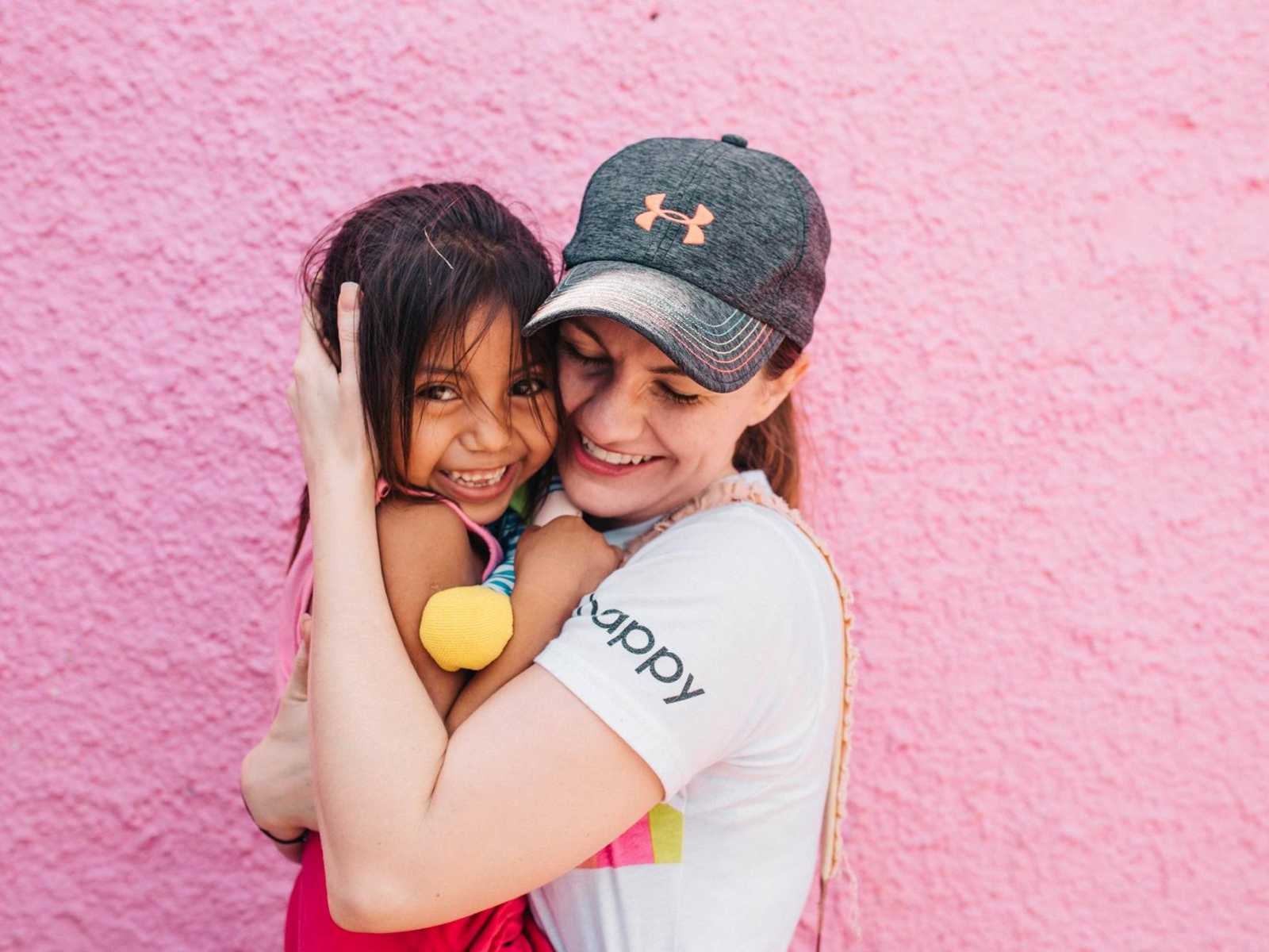 Mother holds adoptive daughter from Mexico close to chest in front of pink wall
