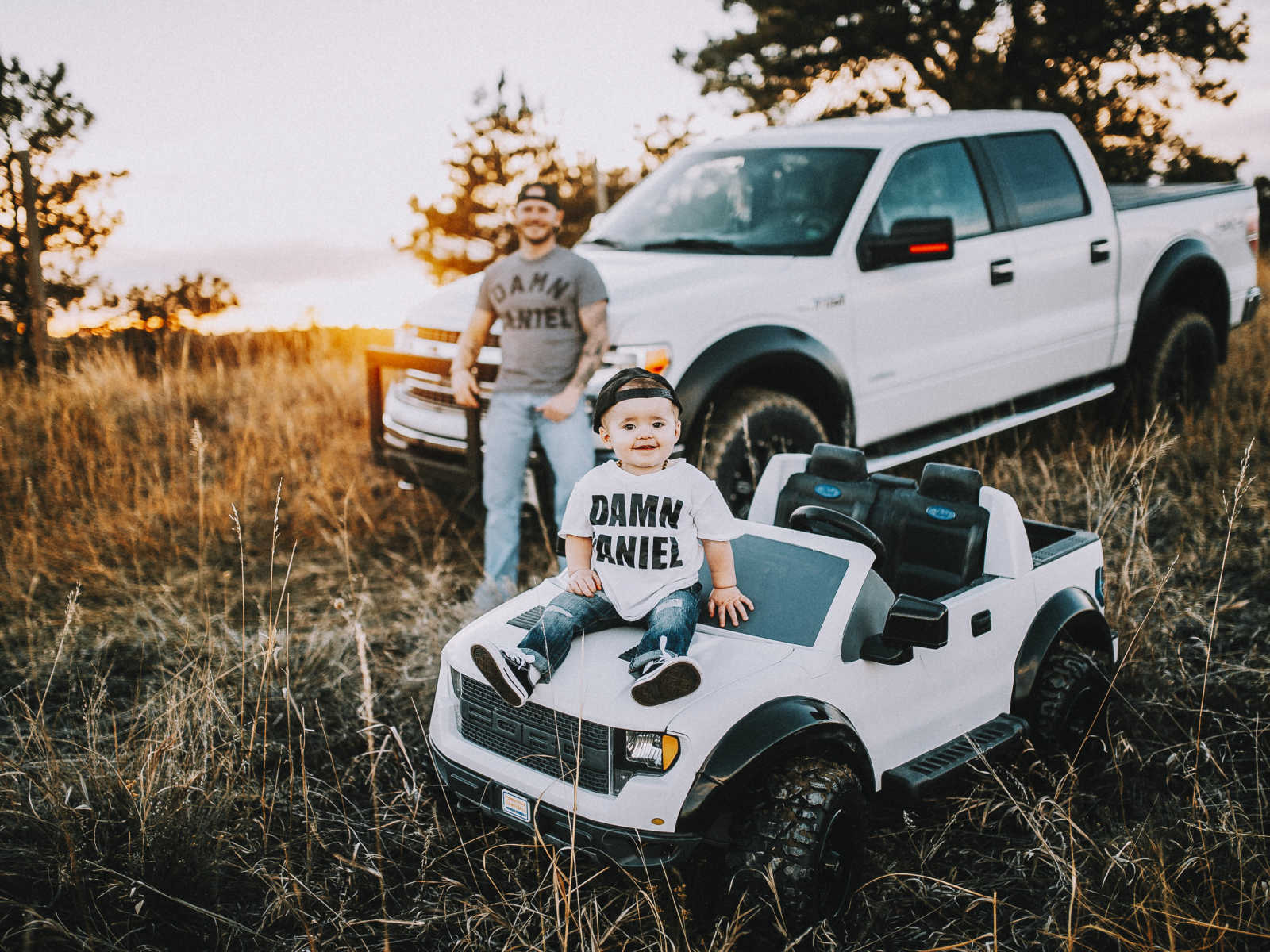 Toddler sits on hood of toy truck with dad wearing the same outfit in background leaning on his truck