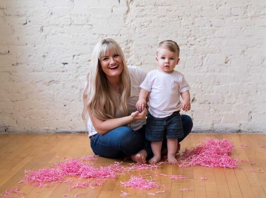 Mother sits on floor with pink confetti next to toddler son who is standing
