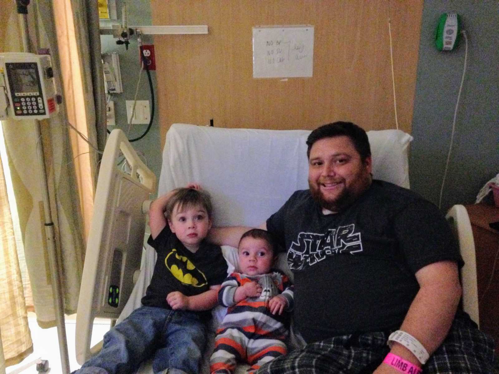 Man who needs kidney transplant sitting in hospital bed with his toddler and baby son next to him