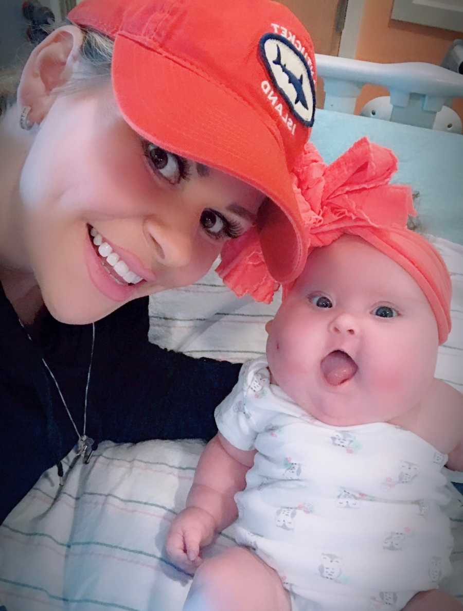 Mother smiles in selfie with baby daughter in hospital bed with large orange bow on her head